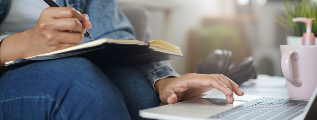 A person taking notes in a notebook while using a laptop